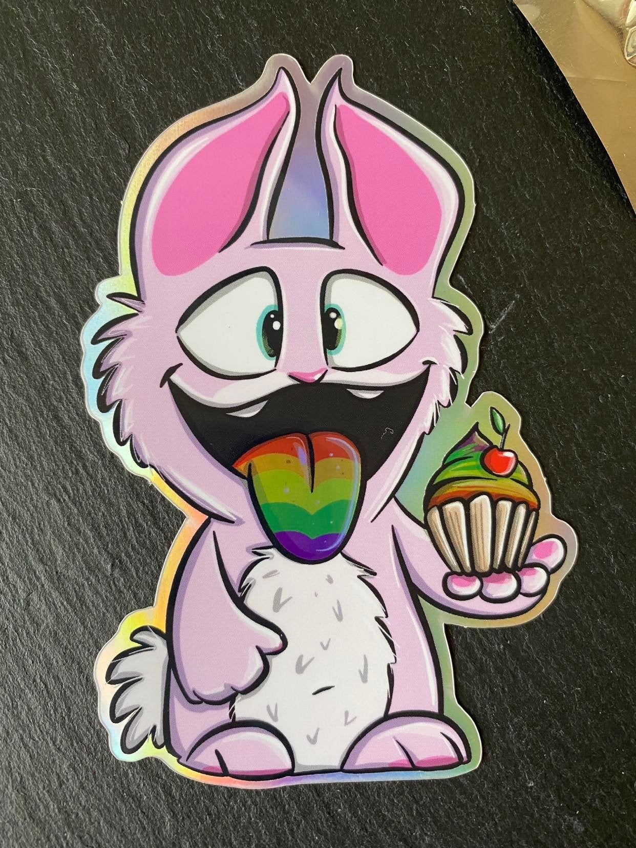A holographic sticker featuring a cartoon design of a character with pink and white features, holding a cupcake.